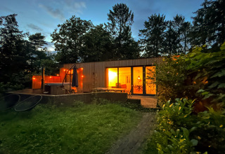 Aftenfoto af Wellnes suite, en lodge i Holland, med oplyst interiør og jacuzziterrasse omgivet af skov.