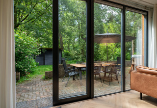 Udsigt fra stue til terrasse med spisebord, stole og parasol i Wellnes suite i Holland, omgivet af skov.