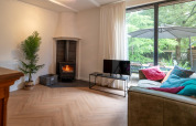 Woonkamer in Forest Suite, Nederland, met houtkachel, sofa, kleurrijke kussens en tuinzicht.