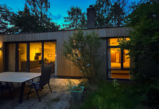 Aftenfoto af Forest suite-lodge i Holland med oplyste vinduer, terrasse og omgivet af grønne træer.