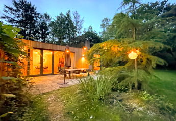 Cozy Forest suite lodge in the Netherlands with glowing lights, patio furniture, and lush greenery at dusk.