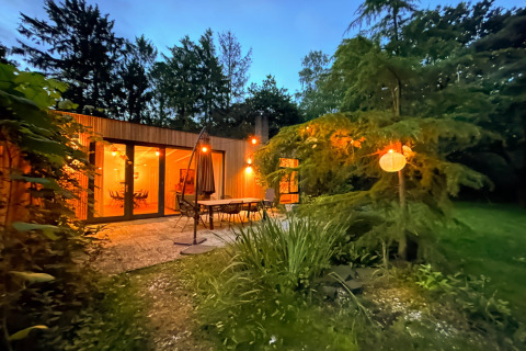 Cozy Forest suite lodge in the Netherlands with glowing lights, patio furniture, and lush greenery at dusk.