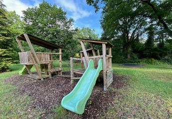 Playground with wooden playhouse, slide, and swings at Forest Suite lodge in the Netherlands, surrounded by trees.