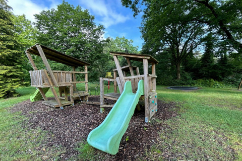 Speelterrein met houten speelhuis, glijbaan en schommels bij Forest Suite lodge in Nederland, omringd door bomen.