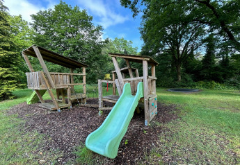 Speelterrein met houten speelhuis, glijbaan en schommels bij Forest Suite lodge in Nederland, omringd door bomen.