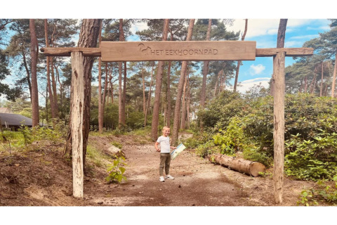 Foto de un niño bajo un cartel de madera 'Het Eekhoornpad' en un sendero de bosque junto a glamping.