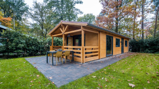 Udvendigt billede af en 4-personers wellness lodge i en skov, med terrasse og bord på flisebelagt område.