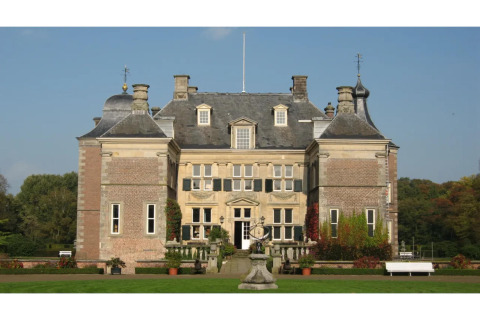 Photo of a large, historic lodge-style mansion with symmetrical architecture, gardens, and lawn in sunlight.