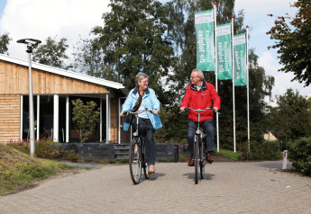 Älteres Paar fährt Fahrrad vor einem Holzhaus im Ferienpark Tolplas 19 in den Niederlanden.