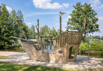Holzspielschiff am Sandstrand eines Sees im Ferienpark Tolplas 19, Niederlande, umgeben von Bäumen.