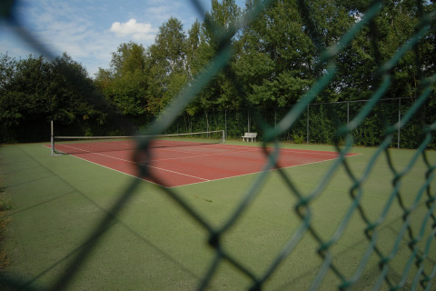 Empty tennis court at Holiday park Tolplas 19, Netherlands, seen through a fence and surrounded by trees.