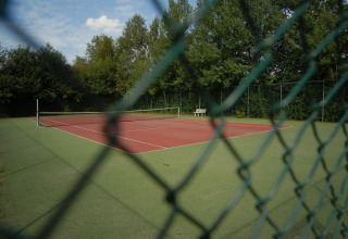 Campo da tennis vuoto al parco vacanze Tolplas 19, Olanda, visto attraverso una rete e circondato da alberi.
