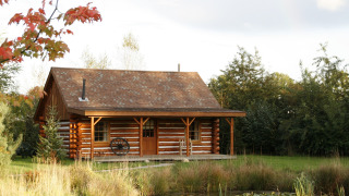 Accogliente chalet chiamato American Log Cabin circondato da alberi, prato e uno stagno di fronte.