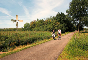 Zwei Menschen fahren Fahrrad auf einer Landstraße neben einer kleinen Windmühle im Tolplas 19, Niederlande.
