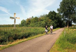 To personer cykler på en landevej ved siden af en lille vindmølle og græs ved Tolplas 19, Holland.