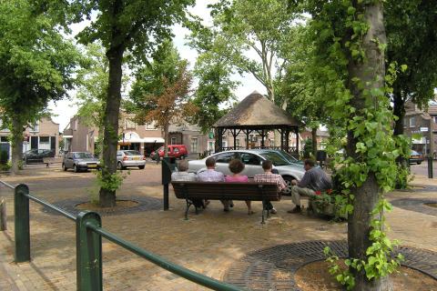 People sit on a bench in a leafy village square with cars and a thatched pavilion at American Log Cabin, Netherlands.
