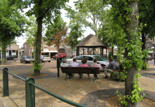 People sit on a bench in a leafy village square with cars and a thatched pavilion at American Log Cabin, Netherlands.