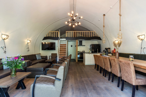 Gezellige lodge woonkamer met gebogen plafond, grote eettafel, zithoek en keuken in Residence De Eese.