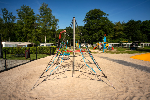 Buitenspeeltuin met klimrekpiramide in zand, omringd door bomen en caravans bij Residence De Eese 22.