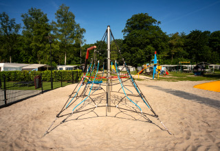 Buitenspeeltuin met klimrekpiramide in zand, omringd door bomen en caravans bij Residence De Eese 22.