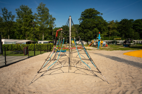 Udendørs legeplads med klatrepyramide i sand, omgivet af træer og campingvogne på Residence De Eese 22.
