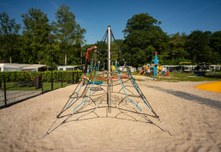 Udendørs legeplads med klatrepyramide i sand, omgivet af træer og campingvogne på Residence De Eese 22.