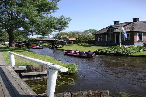 Idyllisk kanal ved Residence De Eese 22 i Holland med mennesker i både og et charmerende hus med flag.