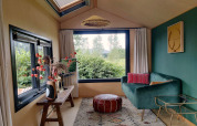 Knusse woonkamer in tiny house in Nederland met groene bank, groot raam, poef en sfeervolle planten.