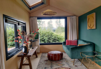 Knusse woonkamer in tiny house in Nederland met groene bank, groot raam, poef en sfeervolle planten.