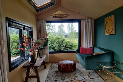 Cozy tiny house living room in the Netherlands with green sofa, large window, pouf, and decorative plants.