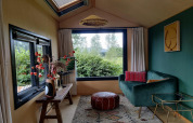 Cozy tiny house living room in the Netherlands with green sofa, large window, pouf, and decorative plants.