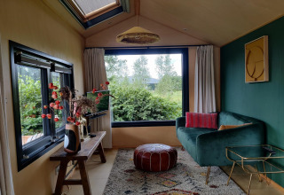 Salon confortable dans une tiny house aux Pays-Bas avec canapé vert, grande fenêtre et plantes décoratives.