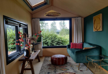 Cozy tiny house living room in the Netherlands with green sofa, large window, pouf, and decorative plants.