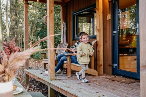 Niño pequeño juega en el porche de una tiny house en el bosque en los Países Bajos con un adulto detrás.