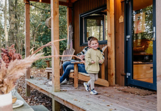 Niño pequeño juega en el porche de una tiny house en el bosque en los Países Bajos con un adulto detrás.