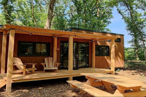 Tiny wooden house with a porch, two Adirondack chairs, and picnic table, set in a forest in the Netherlands.