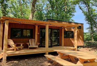 Pequeña casa de madera con porche, dos sillas y mesa de picnic rodeada de bosque en los Países Bajos.