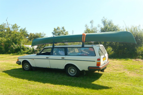 En hvid stationcar med en kano på taget står parkeret på en grøn mark nær glamping-indkvartering.