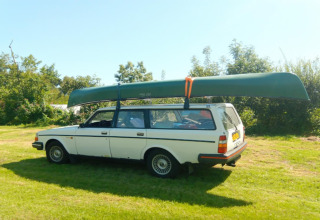 Een witte stationwagen met een kano op het dak staat geparkeerd op gras bij een glamping-accommodatie.