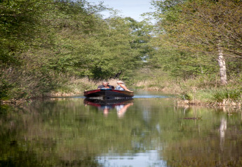 Twee mensen varen in een klein bootje over een smal kanaal, omringd door bomen bij een tiny house in Nederland.