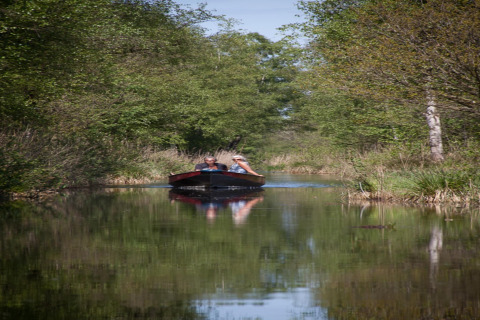 Dos personas navegan en una pequeña barca por un estrecho canal, rodeados de árboles cerca de una tiny house en Holanda.