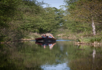 Two people in a small boat travel down a narrow canal surrounded by lush trees near a tiny house in Holland.