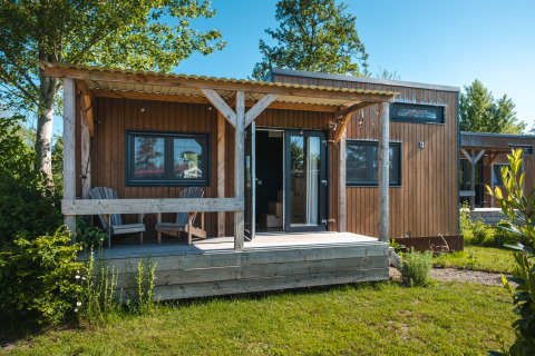 Pequeña casa de madera con porche y dos sillas, Tiny House para 2 personas en un entorno holandés verde.