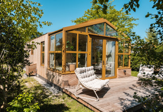 Lodge 'Large Green House 4 persons' nei Paesi Bassi con pareti vetrate, terrazza in legno e sedie da esterno.