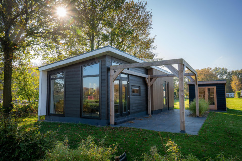 Lodge moderna con grandi finestre e terrazza in legno, circondata da alberi verdi e luce del sole.
