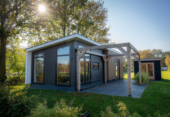 Lodge moderna con grandi finestre e terrazza in legno, circondata da alberi verdi e luce del sole.