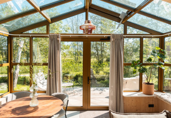 Zonlicht stroomt binnen in een moderne lodge met glazen dak, houten kaders en zicht op de natuur buiten.