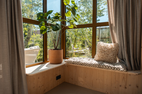 Angolo finestra soleggiato nella lodge «Large Green House» con cuscino, pianta e vista sul verde.