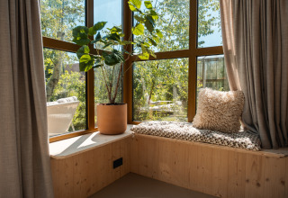 Angolo finestra soleggiato nella lodge «Large Green House» con cuscino, pianta e vista sul verde.