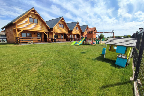 Cabañas de madera en Lazurowy Zakątek con zona infantil y césped verde bajo un cielo azul claro.